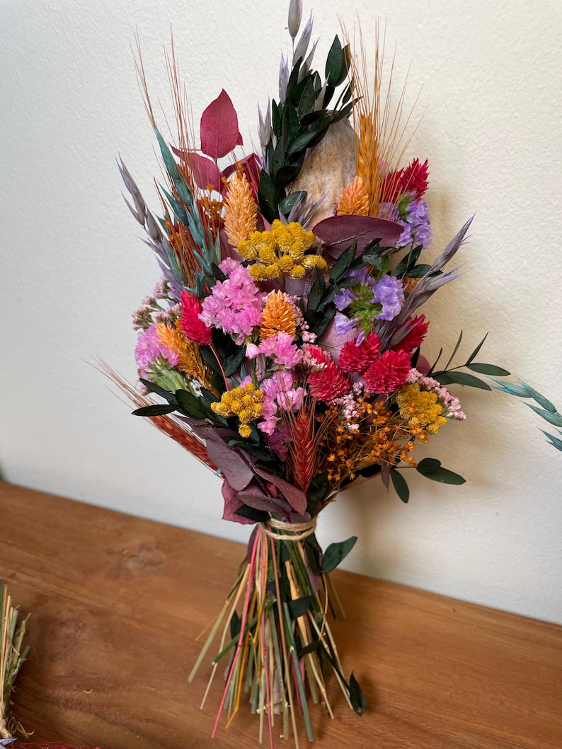 May include: A dried flower bouquet with a variety of colors, including pink, red, yellow, orange, and purple. The bouquet is tied with twine and includes dried grasses and other natural elements.