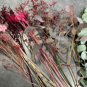 May include: A collection of dried flowers and foliage in shades of pink, red, and green. The arrangement includes eucalyptus, statice, and other textured stems, perfect for crafting or home decor.