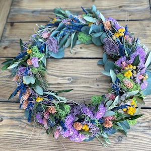 May include: A colourful floral wreath composed of dried flowers in shades of purple, blue, yellow, and pink, with green foliage. The wreath is displayed on a rustic wooden surface, creating a natural and vibrant display.