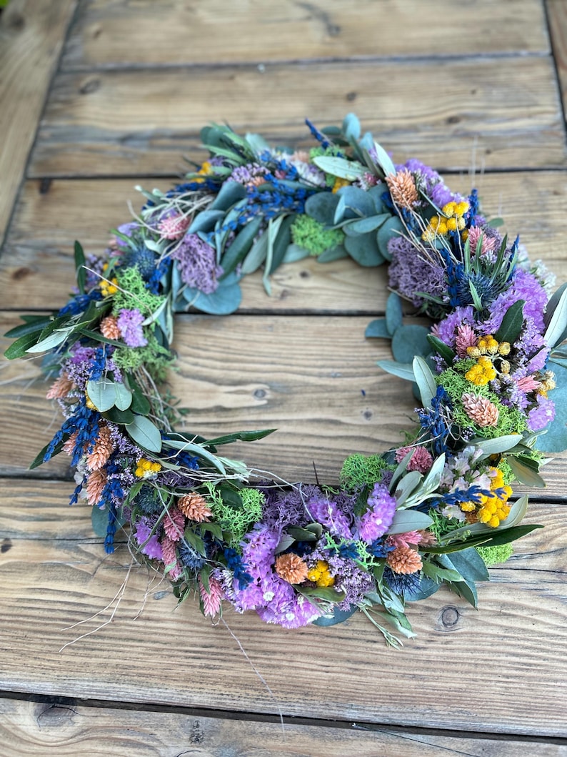 May include: A colourful floral wreath composed of dried flowers in shades of purple, blue, yellow, and pink, with green foliage. The wreath is displayed on a rustic wooden surface, creating a natural and vibrant display.