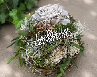 May include: A circular floral arrangement featuring a large, light gray wooden rose at the center, surrounded by green moss, thin twigs, and white lichen. A white cutout sign with the German text IN LIEBEVOLLER ERINNERUNG is placed in front.