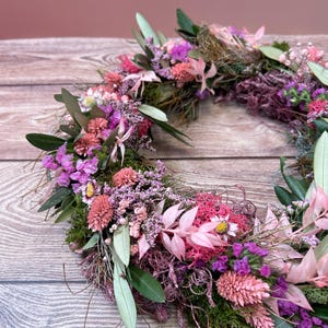 May include: A circular floral wreath made from dried flowers and foliage. The wreath includes pink, purple, and green elements, such as small flowers, leaves, and moss. The arrangement is displayed against a wooden background.