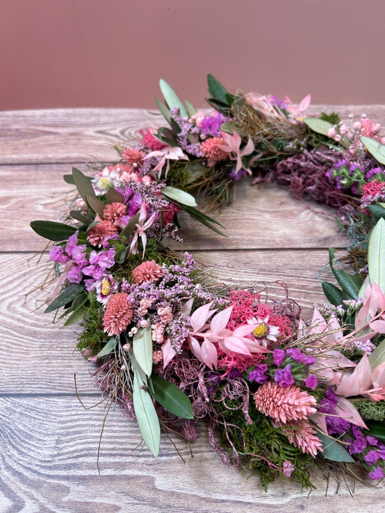 May include: A circular floral wreath made from dried flowers and foliage. The wreath includes pink, purple, and green elements, such as small flowers, leaves, and moss. The arrangement is displayed against a wooden background.