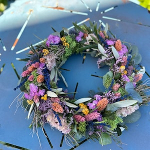 May include: A colourful floral wreath composed of dried flowers in shades of purple, pink, orange, and yellow. The wreath incorporates green foliage and is arranged in a circular form. The wreath is displayed on a blue metal surface.