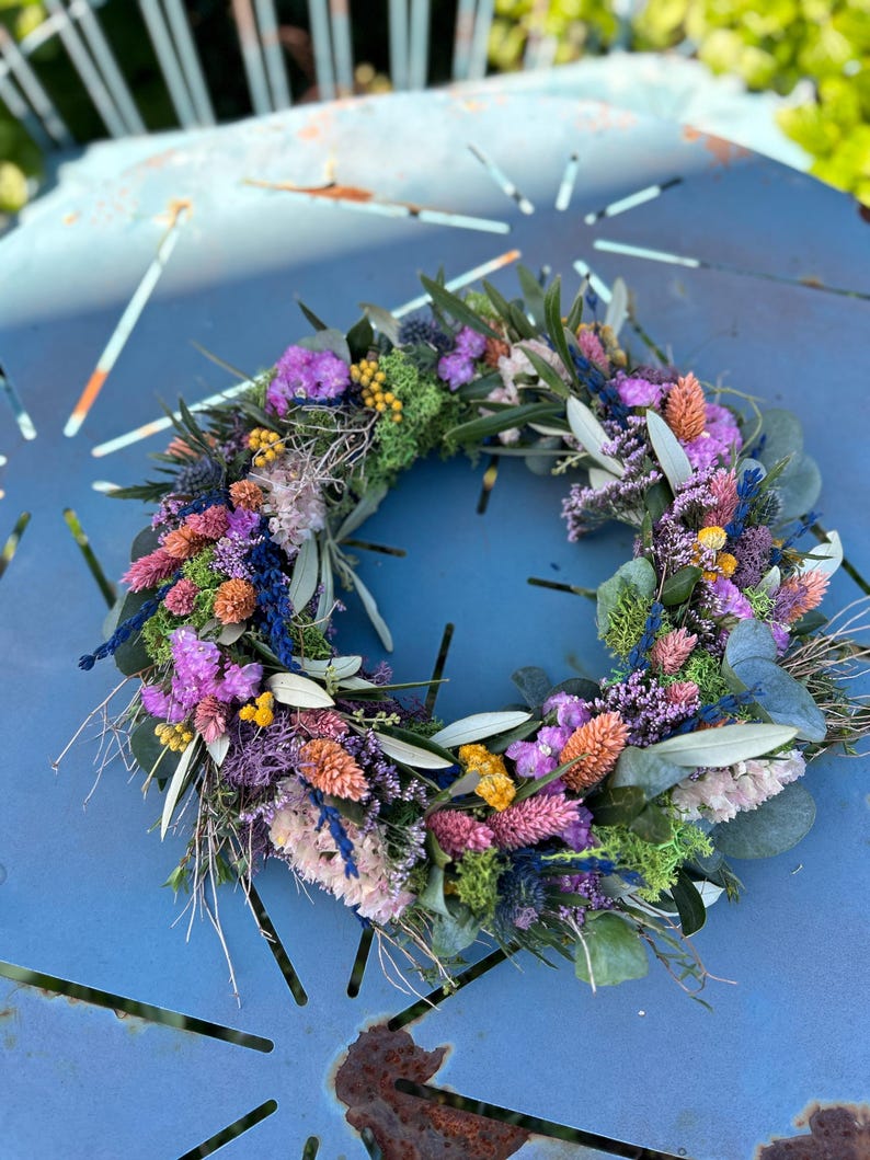 May include: A colourful floral wreath composed of dried flowers in shades of purple, pink, orange, and yellow. The wreath incorporates green foliage and is arranged in a circular form. The wreath is displayed on a blue metal surface.