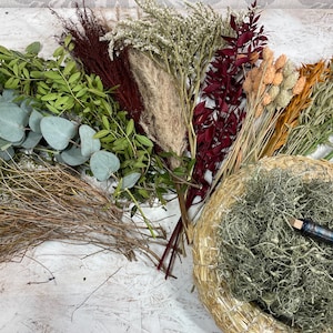 May include: An assortment of dried flowers and greenery, including eucalyptus, pampas grass, and various seed heads. A woven basket filled with dried moss and a pen with a blue plaid pattern are also present. The arrangement features a variety of textures and colors.
