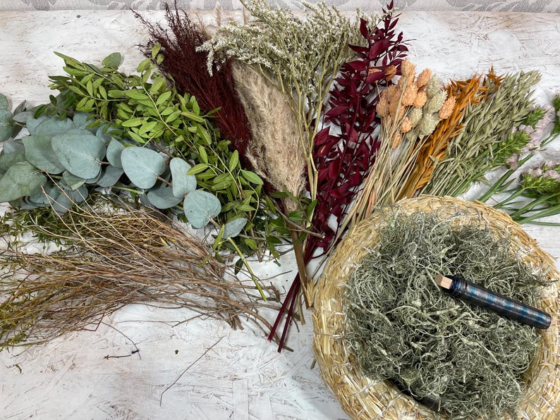 May include: An assortment of dried flowers and greenery, including eucalyptus, pampas grass, and various seed heads. A woven basket filled with dried moss and a pen with a blue plaid pattern are also present. The arrangement features a variety of textures and colors.
