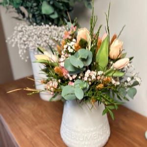 May include: A white ceramic vase filled with a bouquet of flowers, including peach-colored roses, green eucalyptus leaves, and small white and orange blossoms. The vase has embossed text around the base and sits on a wooden surface.