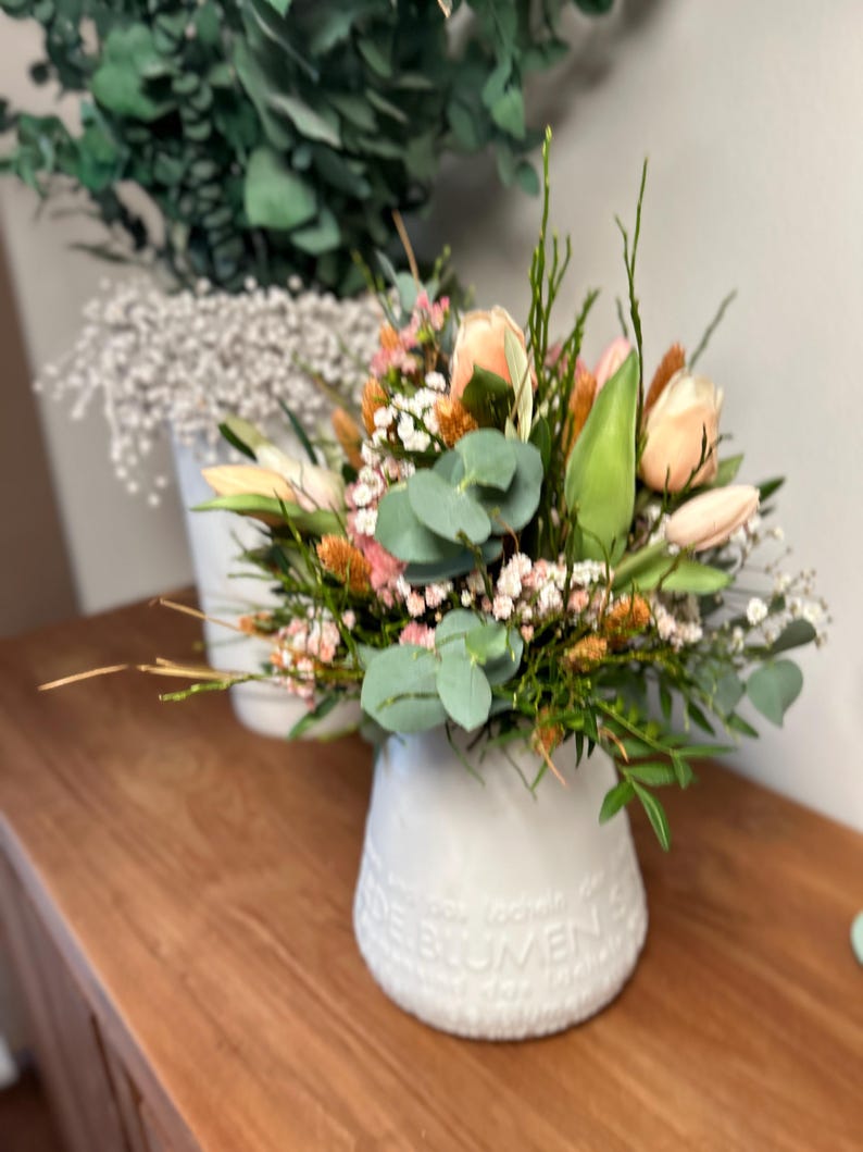 May include: A white ceramic vase filled with a bouquet of flowers, including peach-colored roses, green eucalyptus leaves, and small white and orange blossoms. The vase has embossed text around the base and sits on a wooden surface.