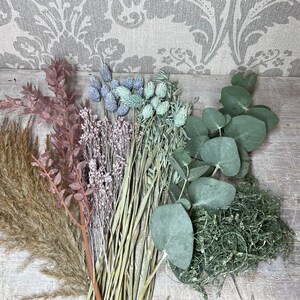 May include: An assortment of dried flowers and greenery, including pampas grass, eucalyptus, and thistle. The color palette includes shades of brown, pink, green, and blue. The arrangement is set against a rustic wooden surface and a patterned backdrop.