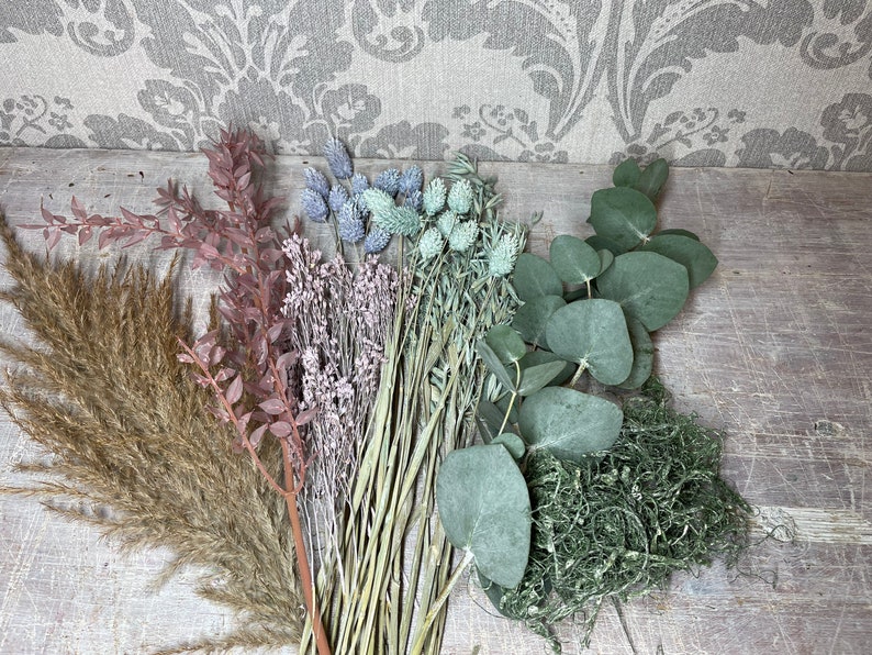 May include: An assortment of dried flowers and greenery, including pampas grass, eucalyptus, and thistle. The color palette includes shades of brown, pink, green, and blue. The arrangement is set against a rustic wooden surface and a patterned backdrop.