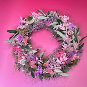 May include: A circular wreath made from dried flowers and foliage. The wreath displays a mix of pink, purple, and green elements, including leaves, small flowers, and textured moss. The background is a solid pink colour.