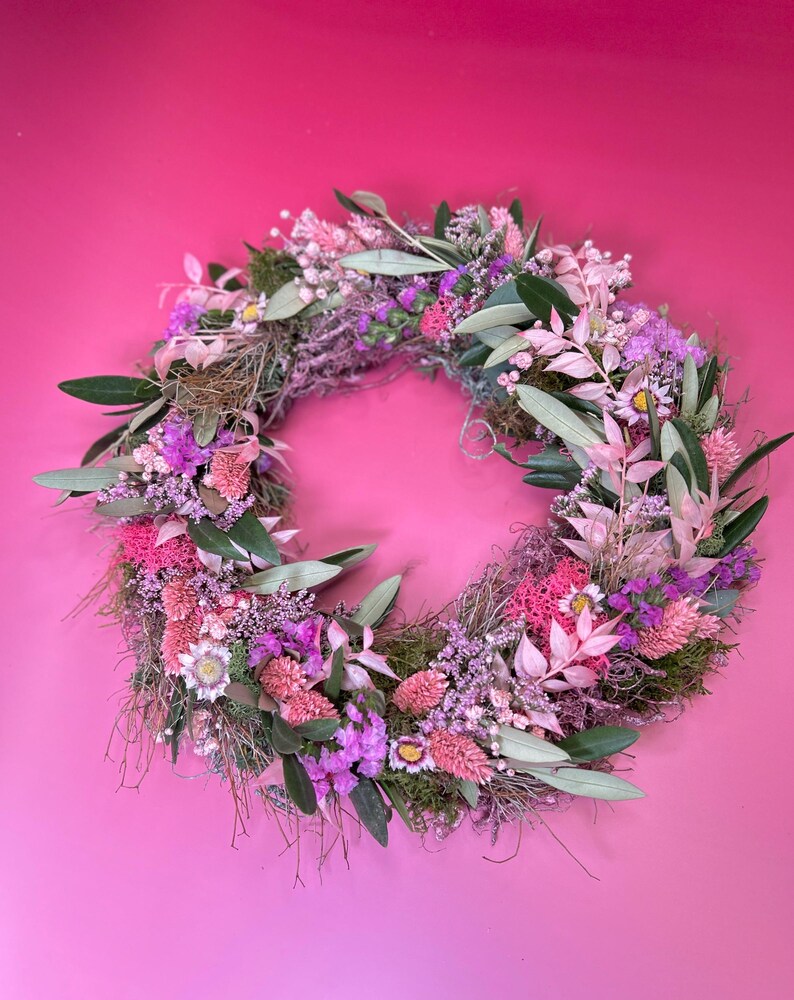 May include: A circular wreath made from dried flowers and foliage. The wreath displays a mix of pink, purple, and green elements, including leaves, small flowers, and textured moss. The background is a solid pink colour.