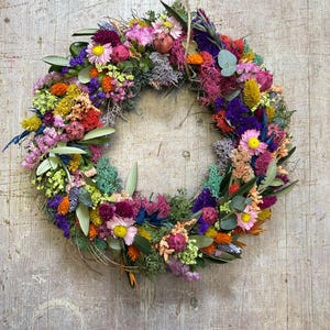 May include: A colourful floral wreath made from dried flowers and foliage, suspended by twine. The wreath features a variety of colours, including pink, yellow, orange, and purple, against a weathered wooden backdrop. The diameter is approximately 30 cm.
