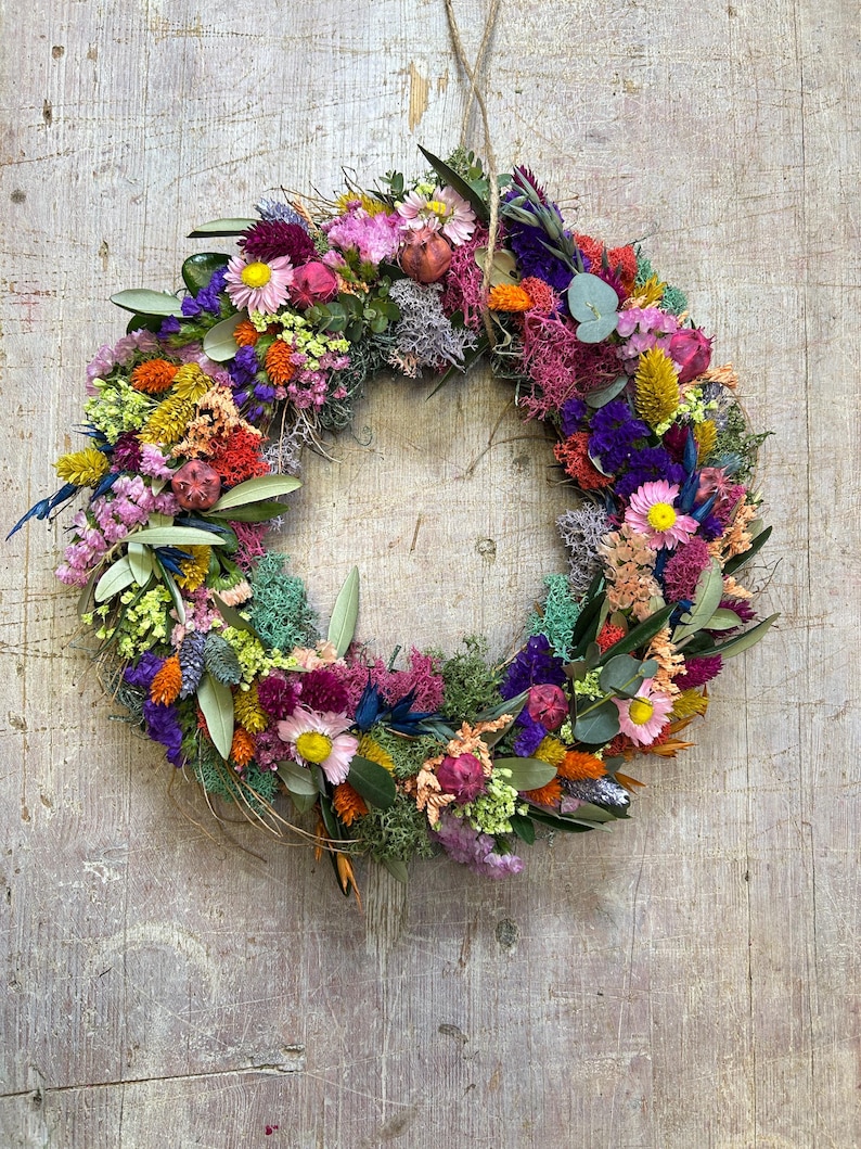 May include: A colourful floral wreath made from dried flowers and foliage, suspended by twine. The wreath features a variety of colours, including pink, yellow, orange, and purple, against a weathered wooden backdrop. The diameter is approximately 30 cm.