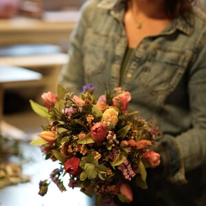 May include: A vibrant bouquet of fresh flowers, featuring tulips in shades of red, pink, and yellow, accented with greenery and small purple blossoms. The arrangement is held by a person wearing a denim shirt.