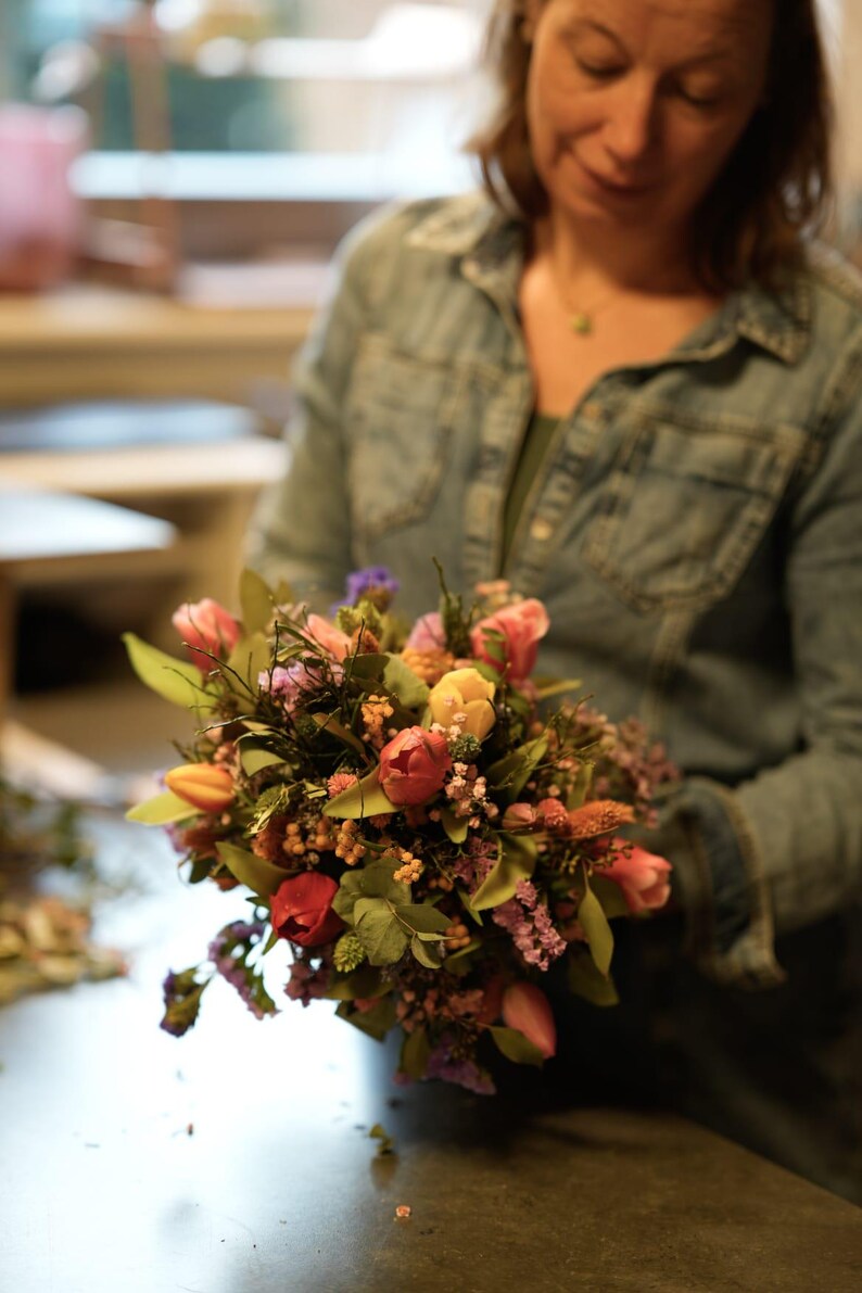 May include: A vibrant bouquet of fresh flowers, featuring tulips in shades of red, pink, and yellow, accented with greenery and small purple blossoms. The arrangement is held by a person wearing a denim shirt.