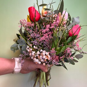 May include: A vibrant bouquet of fresh-cut flowers, featuring bright red, yellow, and pink tulips, accented with delicate pink and white filler flowers and green foliage. The stems are tied together with twine, creating a natural and rustic look.