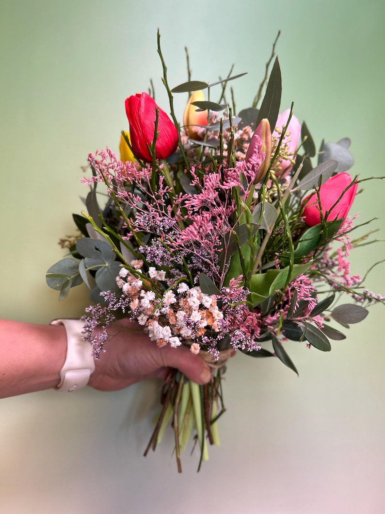 May include: A vibrant bouquet of fresh-cut flowers, featuring bright red, yellow, and pink tulips, accented with delicate pink and white filler flowers and green foliage. The stems are tied together with twine, creating a natural and rustic look.
