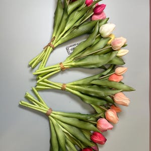 May include: Three bouquets of artificial tulips in various colors, including pink, red, yellow, and peach. The tulips have green stems and leaves, and are tied together with brown twine. The background is a solid gray color.