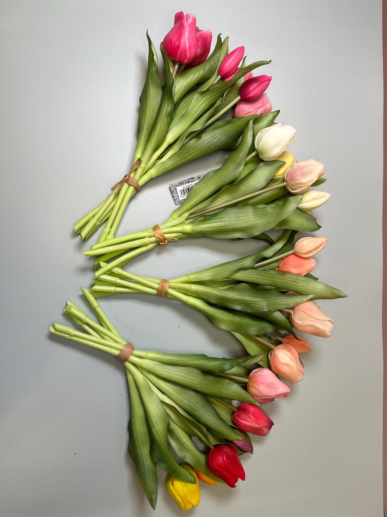 May include: Three bouquets of artificial tulips in various colors, including pink, red, yellow, and peach. The tulips have green stems and leaves, and are tied together with brown twine. The background is a solid gray color.