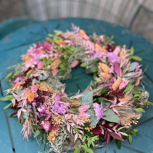 May include: A vibrant floral wreath featuring a variety of dried flowers and greenery. The wreath includes pink, purple, orange, and burgundy blooms, with accents of green foliage and moss. This decorative wreath is perfect for adding a touch of natural beauty to any space.