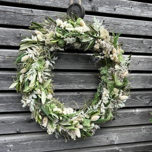 May include: A circular wreath made from dried flowers and foliage in shades of white, green, and beige. It is suspended by a natural rope and a wooden disc against a weathered grey wooden wall.