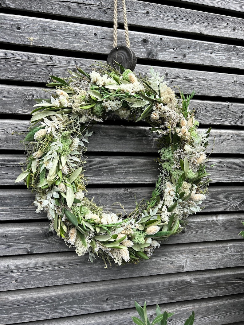 May include: A circular wreath made from dried flowers and foliage in shades of white, green, and beige. It is suspended by a natural rope and a wooden disc against a weathered grey wooden wall.