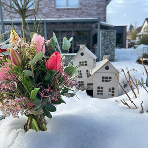 May include: A vibrant bouquet of tulips and other flowers, with pink and red blooms, is tied with twine. Two small, white, paper house decorations sit in the snow. A brick house is in the background.