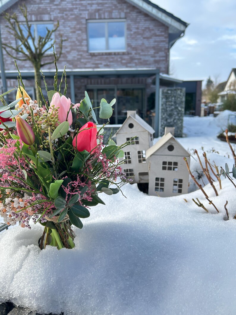 May include: A vibrant bouquet of tulips and other flowers, with pink and red blooms, is tied with twine. Two small, white, paper house decorations sit in the snow. A brick house is in the background.