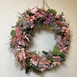 May include: A circular wreath made from dried flowers and foliage. The wreath displays a variety of textures and colours, including pink, green, and brown. The wreath is suspended by a natural fibre cord, and is suitable for home decor.