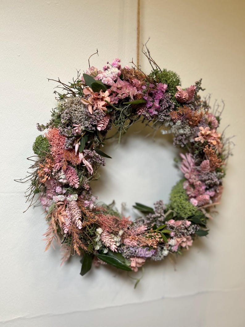 May include: A circular wreath made from dried flowers and foliage. The wreath displays a variety of textures and colours, including pink, green, and brown. The wreath is suspended by a natural fibre cord, and is suitable for home decor.