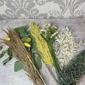May include: An assortment of dried floral stems and greenery, including pampas grass, eucalyptus, and various other dried flowers in shades of brown, green, yellow, and white. The arrangement is set against a light gray background.