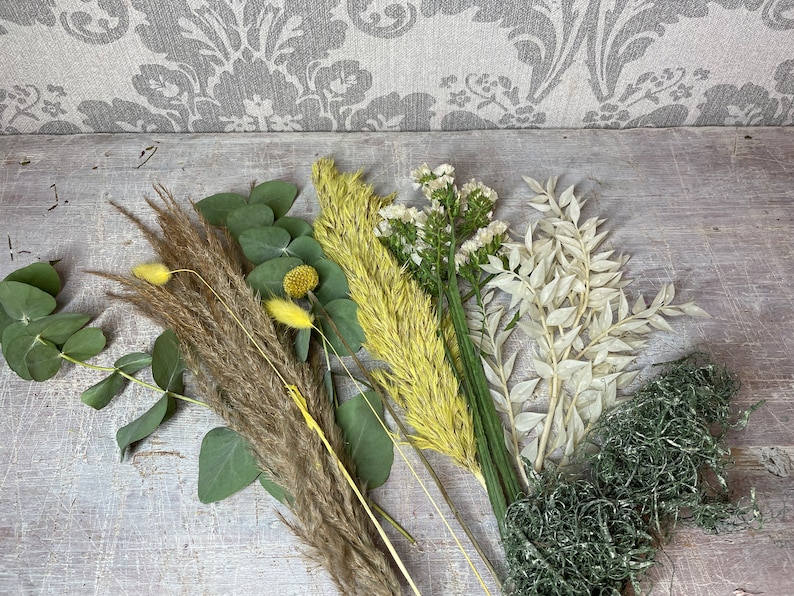 May include: An assortment of dried floral stems and greenery, including pampas grass, eucalyptus, and various other dried flowers in shades of brown, green, yellow, and white. The arrangement is set against a light gray background.