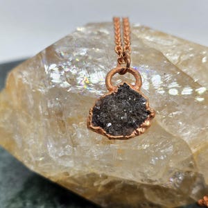 May include: A copper-colored necklace featuring a dark gray, irregularly shaped pendant with sparkling crystals. The pendant is set in a copper frame and hangs from a delicate copper chain. The necklace is displayed on a light-colored, textured stone.