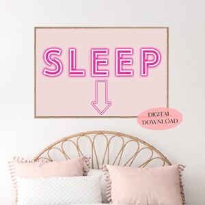 以下が含まれることがあります： ピンクのネオンサインのデジタルダウンロードで、「SLEEP」と書いてあり、下向きの矢印が付いています。サインはピンクの背景にあり、木製のフレームで囲まれています。サインは、ベッド、ナイトスタンド、植物のある寝室の白い壁に掛けられています。