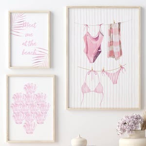 May include: Three framed watercolour prints. One features the text "Meet me at the beach" with pink palm fronds. Another displays pink seashells. The third shows a pink swimsuit and bikini hanging on a clothesline, with a pink and white striped towel.