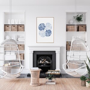 Puede incluir: Una silla colgante de mimbre blanco con una impresi&oacute;n de acuarela azul y blanca de conchas marinas colgando sobre ella. La silla est&aacute; en una sala de estar con una chimenea y una mesa auxiliar de madera.