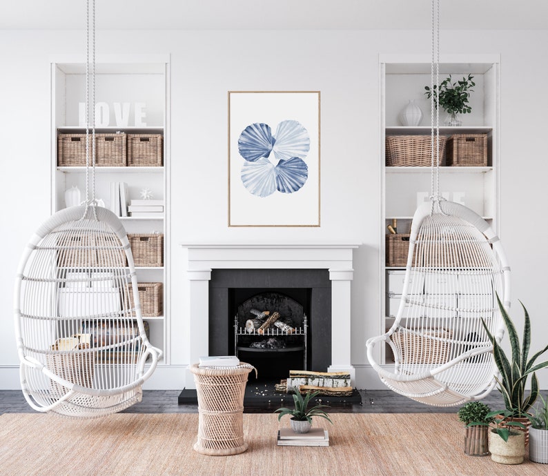 Puede incluir: Una silla colgante de mimbre blanco con una impresi&oacute;n de acuarela azul y blanca de conchas marinas colgando sobre ella. La silla est&aacute; en una sala de estar con una chimenea y una mesa auxiliar de madera.