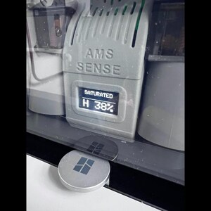 Puede incluir: Primer plano de un dispositivo gris con el texto "AMS SENSE" y una pantalla digital que muestra "SATURADO" y "H 38%". Un bot&oacute;n plateado y circular con un logotipo cuadrado est&aacute; en primer plano. El dispositivo est&aacute; encerrado en una carcasa transparente.