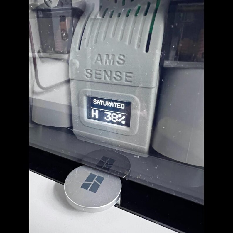 Puede incluir: Primer plano de un dispositivo gris con el texto "AMS SENSE" y una pantalla digital que muestra "SATURADO" y "H 38%". Un bot&oacute;n plateado y circular con un logotipo cuadrado est&aacute; en primer plano. El dispositivo est&aacute; encerrado en una carcasa transparente.