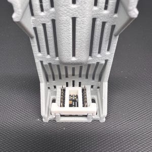 Puede incluir: Un componente gris impreso en 3D con una base rectangular y soportes verticales. Una peque&ntilde;a placa de circuito con texto est&aacute; montada dentro de la estructura. El componente est&aacute; dise&ntilde;ado para electr&oacute;nica o rob&oacute;tica.