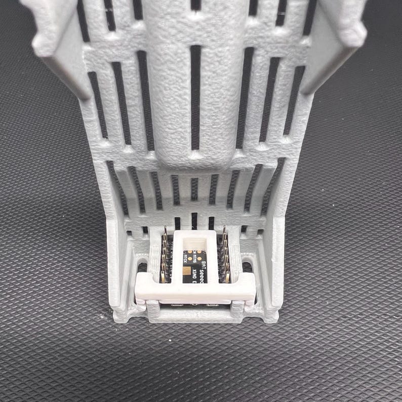 Puede incluir: Un componente gris impreso en 3D con una base rectangular y soportes verticales. Una peque&ntilde;a placa de circuito con texto est&aacute; montada dentro de la estructura. El componente est&aacute; dise&ntilde;ado para electr&oacute;nica o rob&oacute;tica.