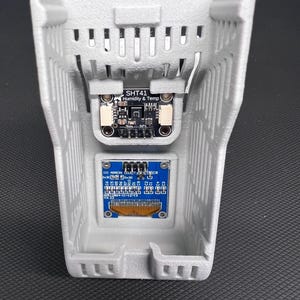 Puede incluir: Una carcasa gris impresa en 3D con ranuras de ventilaci&oacute;n, que contiene dos placas de circuito electr&oacute;nico. La placa superior est&aacute; etiquetada como "SHT41 Humidity & Temp". La placa inferior tiene componentes azules y dorados y texto. La carcasa est&aacute; sobre una superficie oscura.