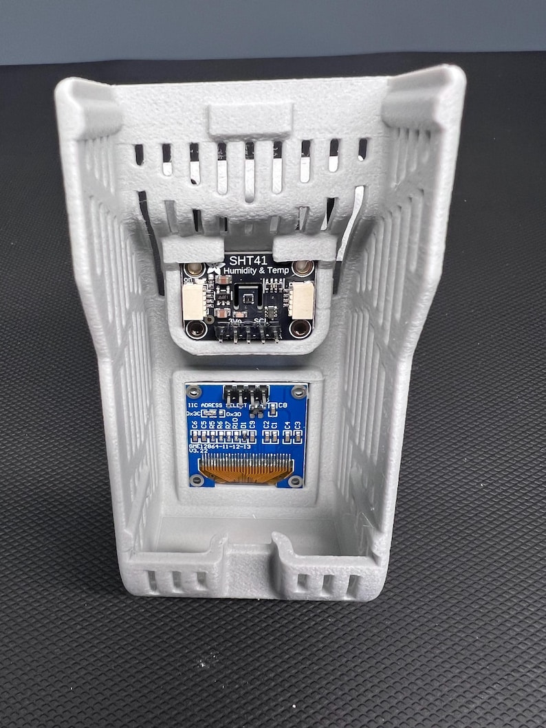 Puede incluir: Una carcasa gris impresa en 3D con ranuras de ventilaci&oacute;n, que contiene dos placas de circuito electr&oacute;nico. La placa superior est&aacute; etiquetada como "SHT41 Humidity & Temp". La placa inferior tiene componentes azules y dorados y texto. La carcasa est&aacute; sobre una superficie oscura.