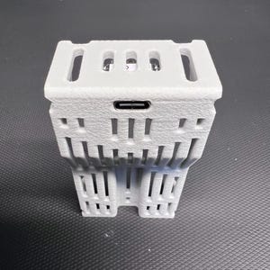 Puede incluir: Una carcasa de dispositivo electr&oacute;nico gris claro, impresa en 3D, con una superficie texturizada. Presenta una forma rectangular con m&uacute;ltiples ranuras rectangulares y verticales. Se ve un puerto USB-C. El dispositivo tiene un dise&ntilde;o geom&eacute;trico &uacute;nico.