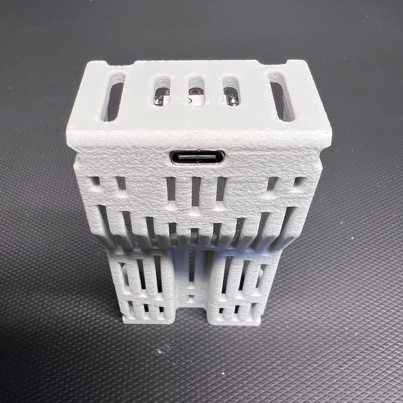 Puede incluir: Una carcasa de dispositivo electr&oacute;nico gris claro, impresa en 3D, con una superficie texturizada. Presenta una forma rectangular con m&uacute;ltiples ranuras rectangulares y verticales. Se ve un puerto USB-C. El dispositivo tiene un dise&ntilde;o geom&eacute;trico &uacute;nico.