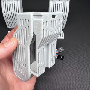 Puede incluir: Un componente gris claro impreso en 3D con forma rectangular y dise&ntilde;o ranurado vertical. El componente est&aacute; sostenido por una mano, con varios componentes similares en el fondo. Tambi&eacute;n se ve un conector multicolor.