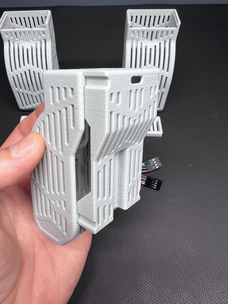 Puede incluir: Un componente gris claro impreso en 3D con forma rectangular y dise&ntilde;o ranurado vertical. El componente est&aacute; sostenido por una mano, con varios componentes similares en el fondo. Tambi&eacute;n se ve un conector multicolor.