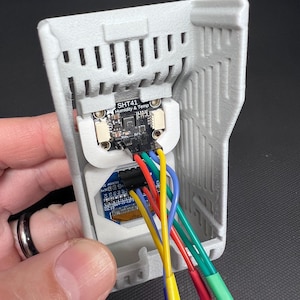 Puede incluir: Una carcasa gris impresa en 3D que contiene una placa de circuito electr&oacute;nico con el texto "SHT41 Humidity & Temp". La placa est&aacute; conectada a cables multicolores, incluyendo rojo, amarillo, verde y azul. El dispositivo se sostiene en una mano.